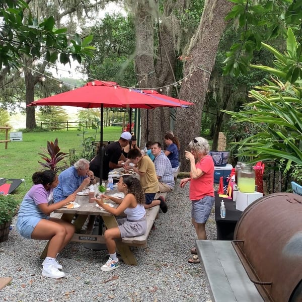 Patrons enjoying a meal at Munchies Live BBQ, seated at picnic tables under red umbrellas outdoors.