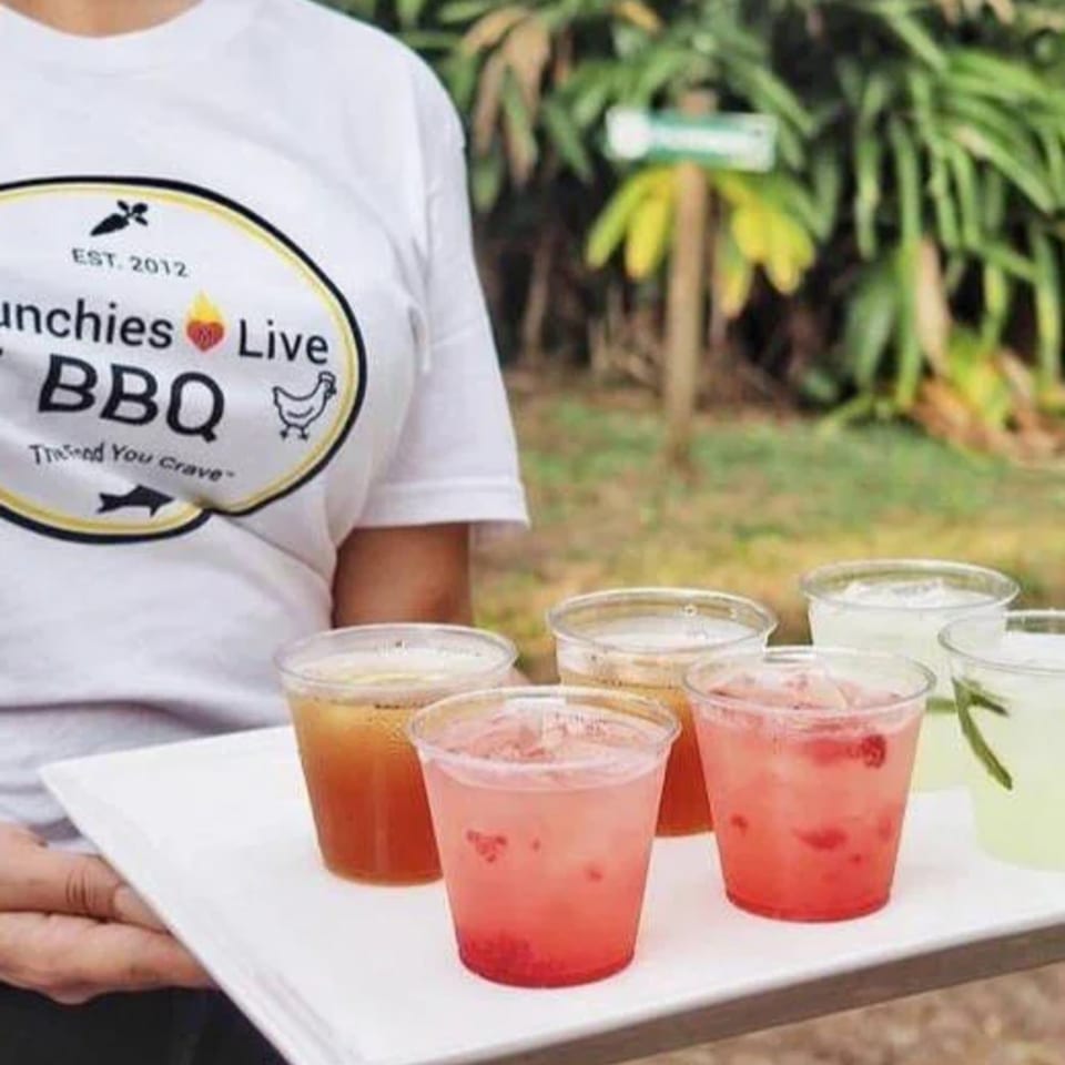 A person wearing a Munchies Live BBQ shirt holds a tray of colorful drinks outdoors.