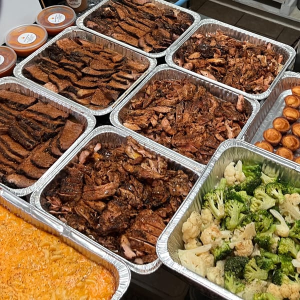 Multiple aluminum trays filled with sliced meats, macaroni and cheese, broccoli, and small pastries.