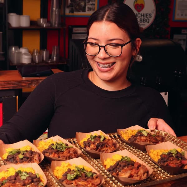 A smiling woman in glasses holds a tray of Munchies Live BBQ pulled pork, rice, and beans.