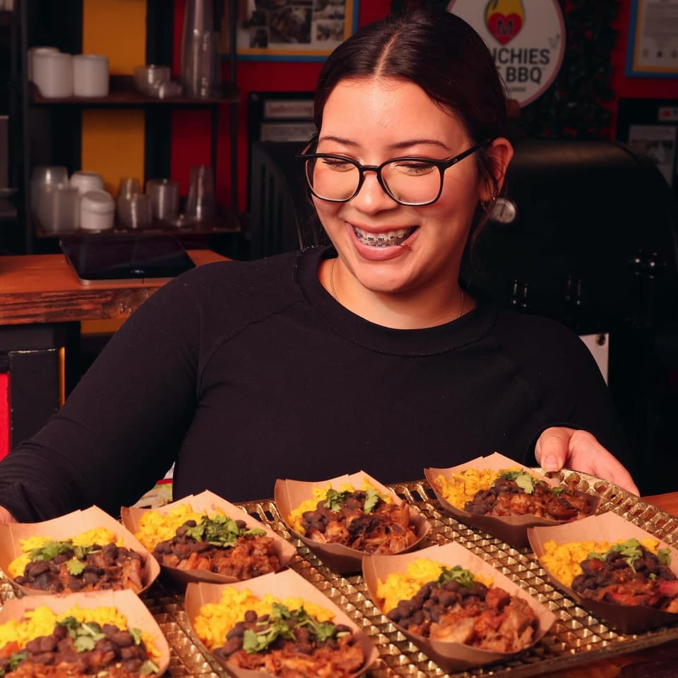 A smiling woman in glasses holds a tray of Munchies Live BBQ pulled pork, rice, and beans.