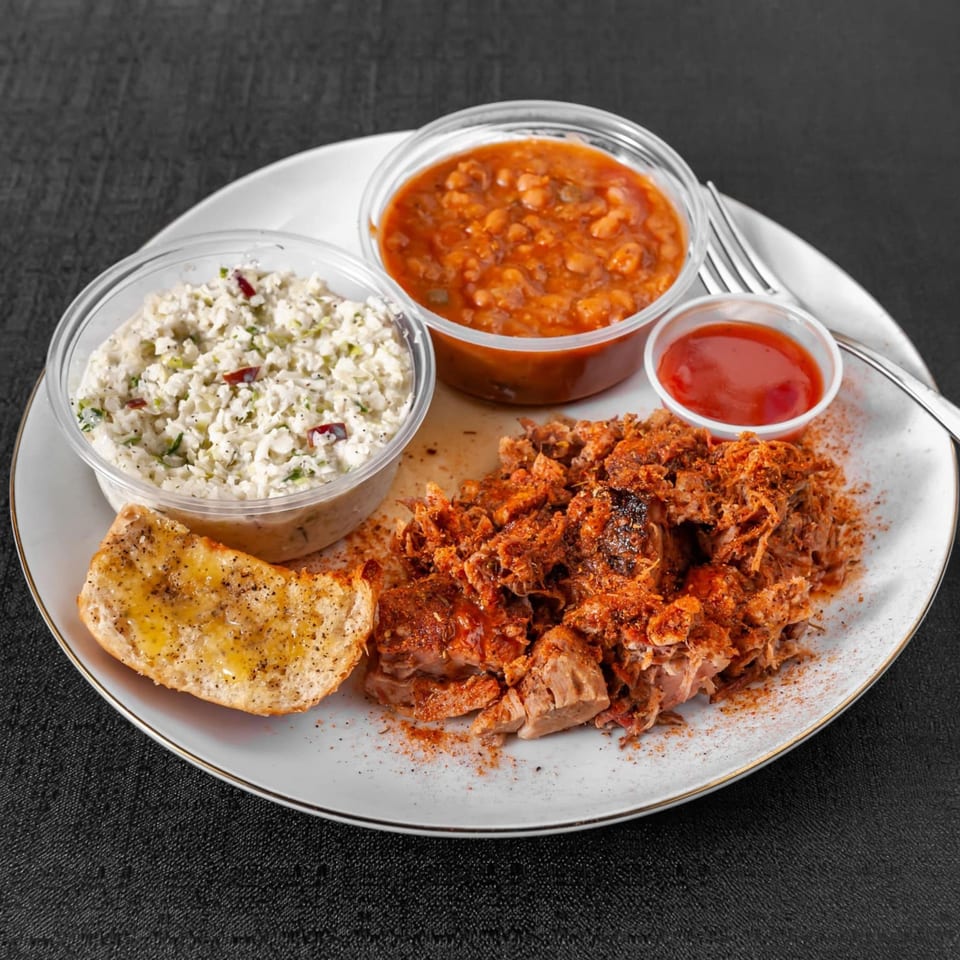 A BBQ plate with pulled pork, baked beans, coleslaw, garlic bread, and a side of sauce.