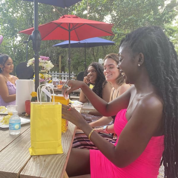 Four women enjoying an outdoor meal at Munchies Live BBQ, with one opening a yellow gift bag.