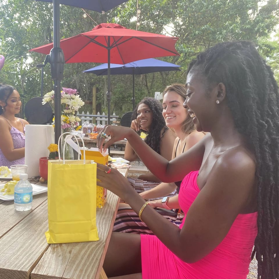 Four women enjoying an outdoor meal at Munchies Live BBQ, with one opening a yellow gift bag.