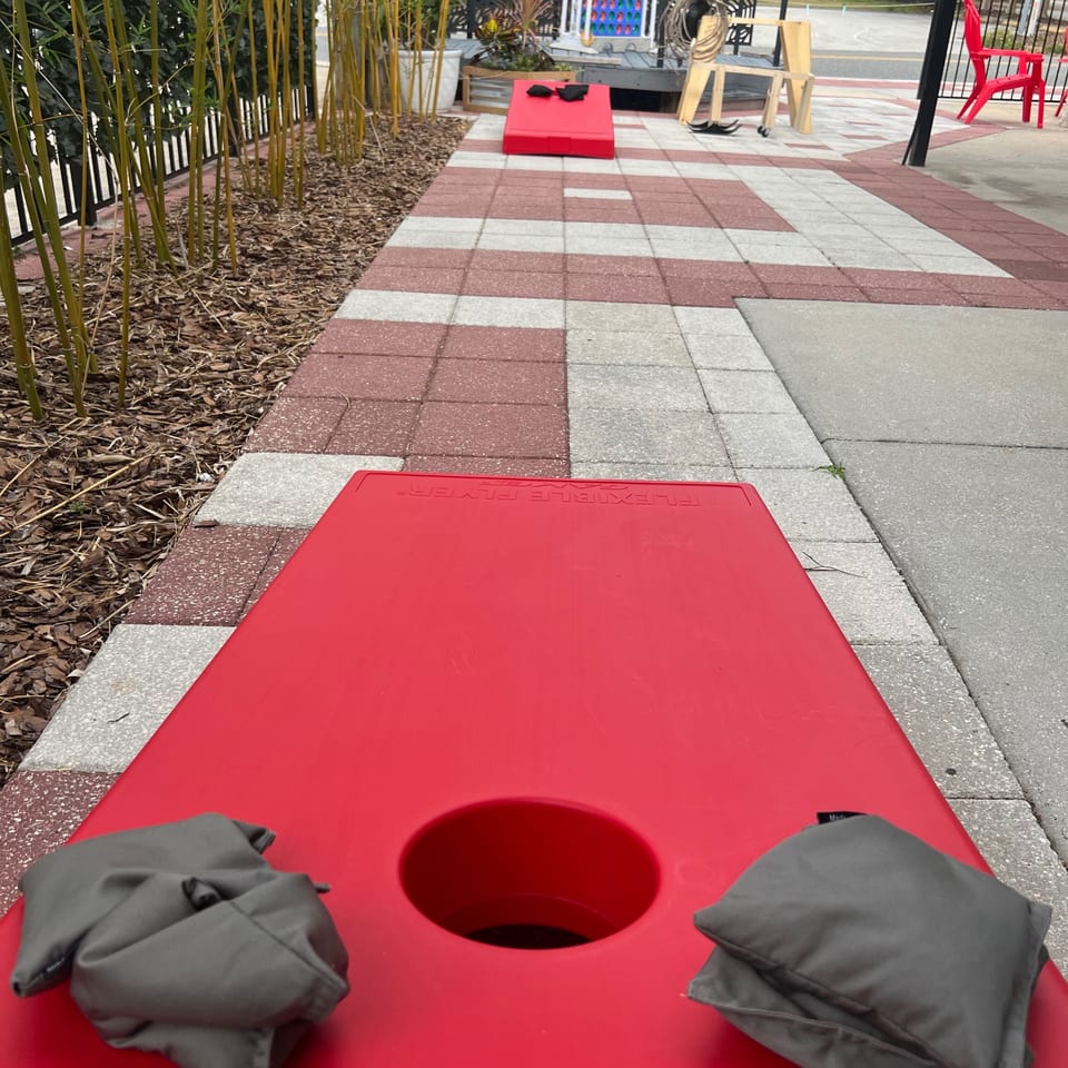 Two red cornhole boards with beanbags on a patio with bamboo and red chairs.